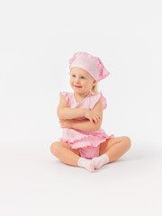 Satisfied, happy little girl 2 years old in a pink dress and a scarf sits on the floor, smiles playfully and hugs herself with her arms.