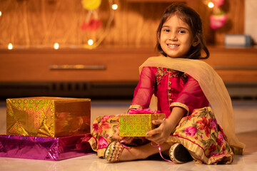 Happy little indian girl kid holding gift boxes in hand celebrating Diwali while sitting on floor,...