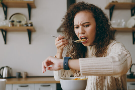 Young Sad Late Woman Wears Casual Clothes Sweater Eat Breakfast Muesli Cereals With Milk Fruit In Bowl Look At Smart Watch Sit At Table In Light Kitchen At Home Alone. Lifestyle Cooking Food Concept.