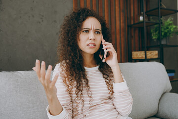 Young sad upset woman wearing casual clothes sits on grey sofa couch talking speak on mobile cell...