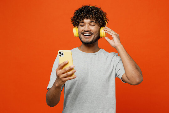 Young smiling cheerful happy Indian man he wears t-shirt casual clothes listen to music in headphones use mobile cell phone isolated on orange red color background studio portrait. Lifestyle concept.