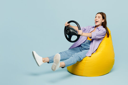 Full body surprised fun excited young happy woman she wears purple shirt yellow t-shirt casual clothes sit in bag chair hold steering wheel driving car isolated on plain pastel light blue background.