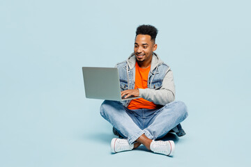 Full body young IT man of African American ethnicity wear denim jacket orange t-shirt sit hold use work on laptop pc computer isolated on plain pastel light blue background studio. Lifestyle concept .