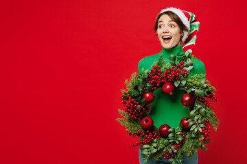 Young amazed woman wear green turtleneck Santa hat posing hold in hand decorative wreath look aside...