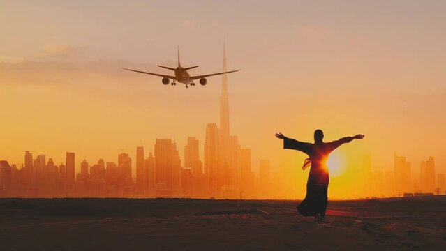 Back view of Arab woman rises her hands on the sun set. Passengers Airplane preparing for landing at Dubai International airport. Dubai city on the background.