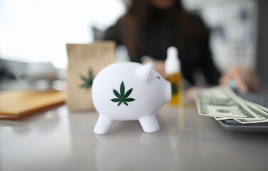 Close-up of piggy bank with green sign of cannabis. Paper package and bottle of cannabinoid oil...