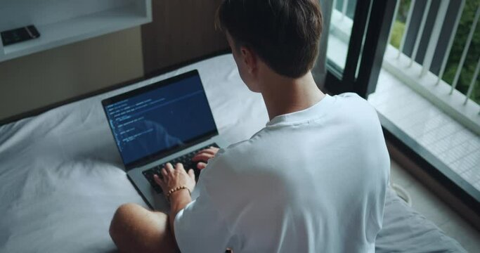 Rear view man freelancer programmer sitting on bed in bedroom and working on laptop computer. Freelance man writing program code using laptop, working remotely from home. Freelance concept