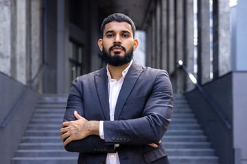 Portrait of a young Muslim male lawyer standing in a business suit outside a courthouse and looking confidently into the camera with his arms crossed