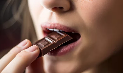 Fotobehang Beautiful young woman eating dark chocolate. close up of mouth,lips and teeth. Beauty model girl enjoying her chocolate bar, close eyes. Pretty brunette female biting chocolate bar over brown  © annebel146
