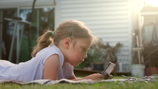 Close up view of Carefree girl is playing on the tablet. Lies on the grass near the house, the sun illuminates her hair. 