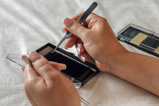 Hands of female lashmaker holding tweezers and carefully taking artificial eyelash from palette lying on white sheet.