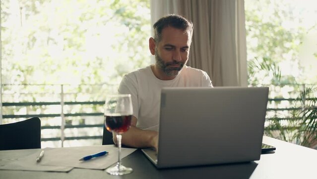 Video of handsome  man working with laptop while drinking a glass of wine in the kitchen at home