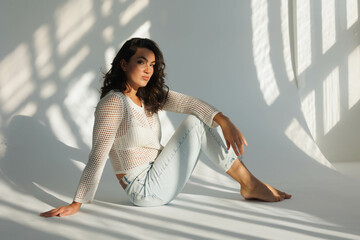 Beautiful brunette woman in white net top and blue jeans, sitting. Girl smile, happy. Portrait of young pretty woman. Complicated sunlight with shadows. White background. Light from window, portrait.