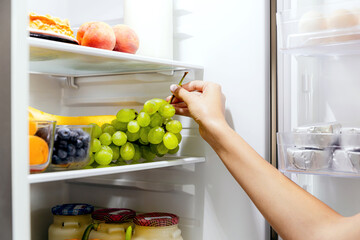 Woman hand taking or picks up bunch of grapes out of open refrigerator or fridge
