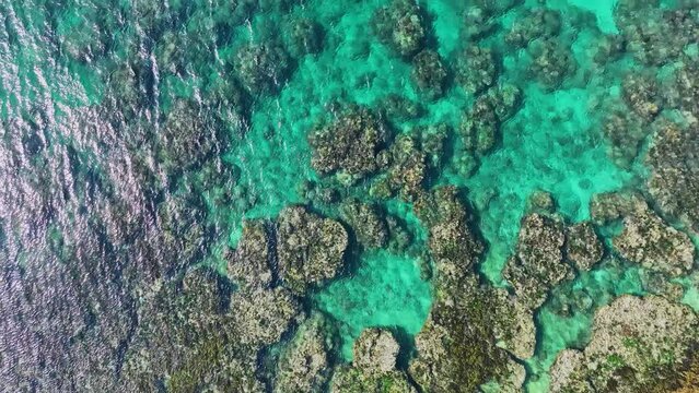 Top view of blue-green sea waves reaching the shore, with coral reefs and rocks, captured in 4K drone view