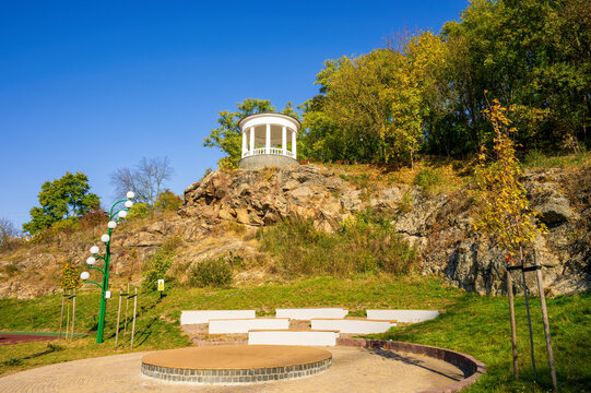 A picturesque view of a city park in Zhytomyr, Ukraine, featuring a white gazebo perched on a rocky cliff amidst lush greenery