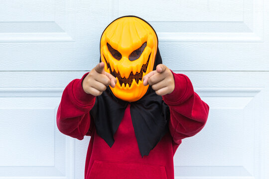 A child girl wearing a scary Halloween pumpkin mask, pointing two fingers at the camera, over a white garage door. Concept of celebration, costume, carnival, terror, fear and autumn.