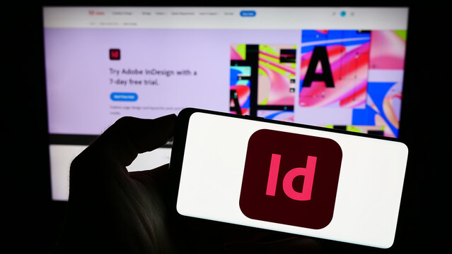 Stuttgart, Germany - 09-23-2023: Person holding mobile phone with logo of desktop publishing software Adobe InDesign on screen in front of web page. Focus on phone display.