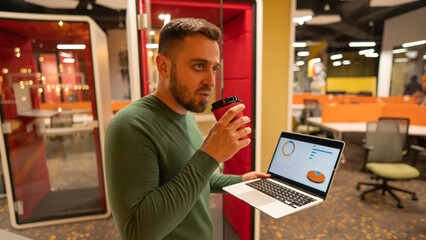 Fototapeta na wymiar Bearded man holding laptop and drinking coffee in open space office.