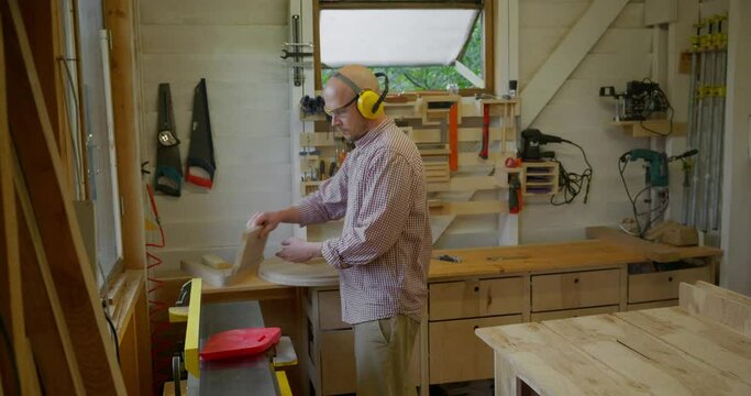 The general plan, a young bald man woodworker working in his small workshop on a jointing machine in front of the window. A carpenter in safety glasses and headphones is whittling a board on a jointer
