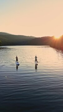 Aerial top down view of two men silhouette standing on stand up paddle board doing sup water sport activity on a mountain lake at sunset. Healthy lifestyle outdoor activity 