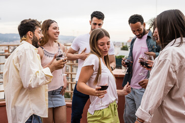 Happy young group of friends dancing in a rooftop party holding drinks.Diverse cheerful people having fun in a terrace drinking wine and listening music.