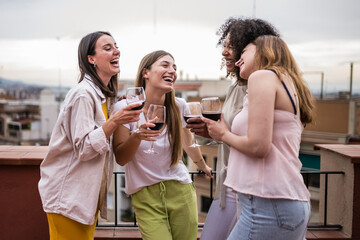 Diverse group of female friends having fun while drinking wine in a rooftop party. Carefree and happy young women laughing standing in a terrace holding glasses.