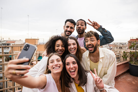 Diverse joyful group of carefree friends having fun and taking a selfie in a rooftop party. Multiracial beautiful young happy people laughing while taking a picture in a terrace.