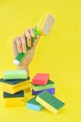 A wooden hand holds a sponge for washing dishes, next to it there is a pile of multi-colored sponges.  Yellow background.  Free space for text.