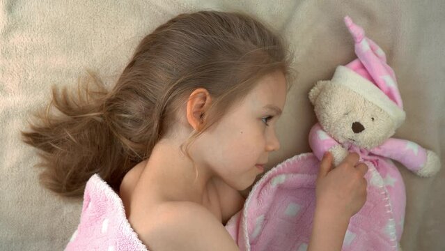 Close up of little girl sleeps at home in bed with toy and smiles. Child in dream hugs teddy bear. Healthy sleep concept