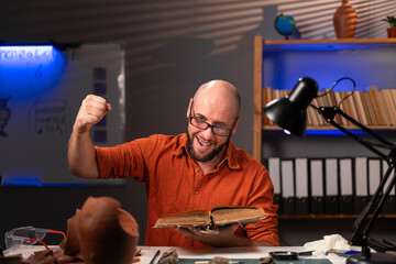 Happy archaeologist working in office late at night reading old book finding solution to problem