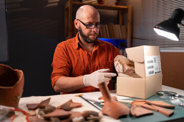 Male archaeologist in glasses working late at night in the office preparing antiques for shipment...
