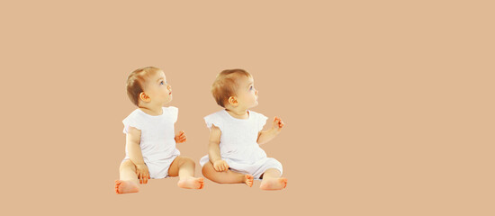 Two twin babies sitting on the floor and looking up together on beige studio background, blank copy space for advertising text