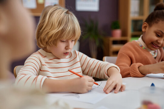 Portrait of cute blonde boy coloring pictures at table in arts and crafts class in preschool, copy space