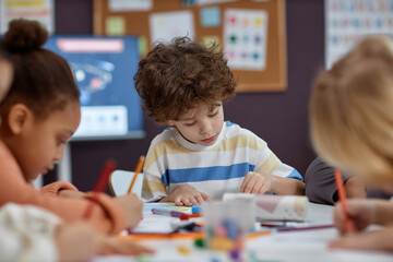 Portrait of cute boy coloring pictures at table with group of children in arts and crafts class in preschool, copy space