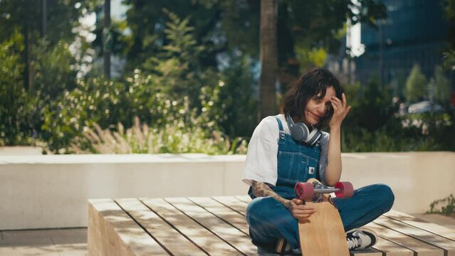 Woman in denim overalls sitting on wooden bench with skateboard, listen music in headphones, downtown city street, relaxed female skateboarder enjoys summer sunlight, urban style, modern architecture