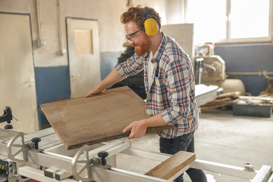Smiling carpenter putting wooden board on circular saw at carpentry manufacturing 