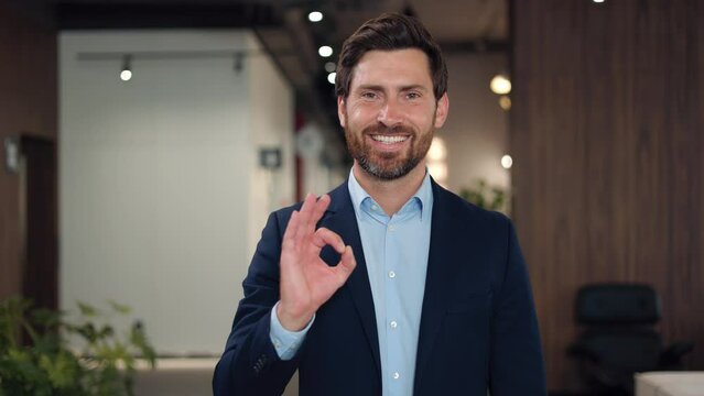 Portrait of handsome businessman showing ok gesture with fingers while expressing positive emotion at career space. Caucasian male in formal suit making hand sign to agree with something indoors.