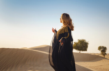 Desert adventure. Young arabian Woman posing in traditional Emirati dress abaya in sanddunes of UAE desert at sunset. The Dubai Desert Conservation Reserve, United Arab Emirates. - obrazy, fototapety, plakaty