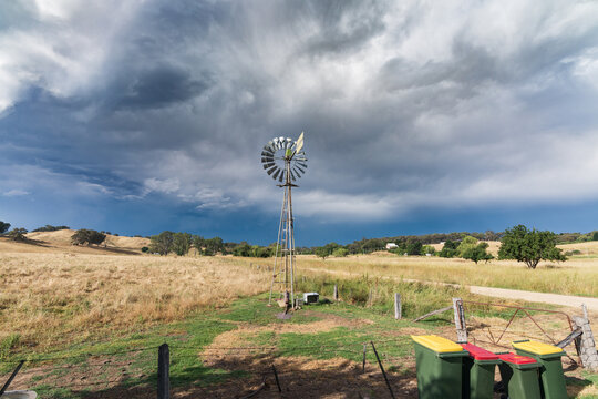 A row of rubbish bins and a still windmill under a dramatic dark stormy sky over dry farmland