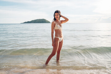 Beautiful caucasian woman in white bikini on tropical beach. Portrait of dark skinned woman smiling at sea. Brunette tanned girl in swimwear enjoying and walking on beach.