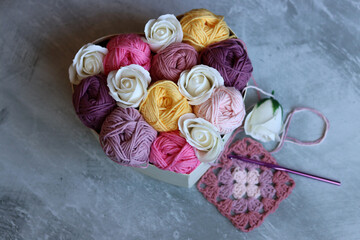 Multicolored balls of cotton yarn and white roses in a basket on a gray background with space for text. Handmade gift box with organic thread. Creative gift for crochet lover. 
