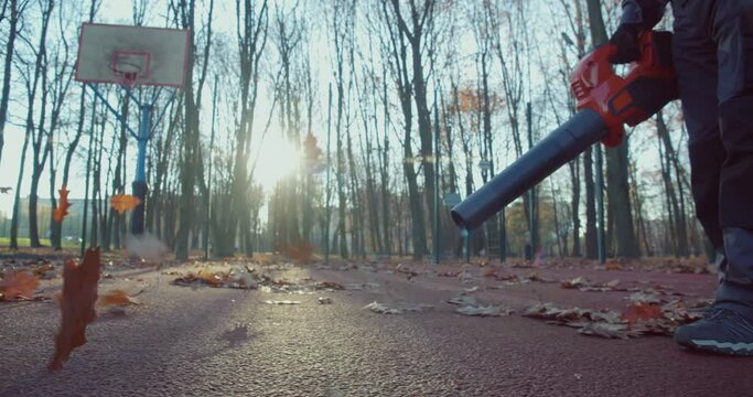 Close up of caucasian man in working clothes using leaf blower for cleaning city park during autumn time. Concept of people and maintenance.