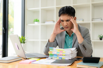Stressed businessman sitting at workplace and touching his headYoung asian exhausted businesssman with messy desk and stack of papers, working busy, overwork.