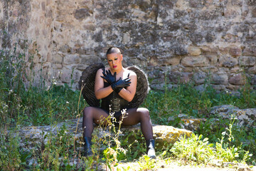 Young non-binary Latin person, wearing black make-up and black angel wings, sitting on a rock and...