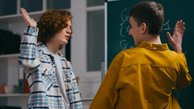 Teen boy helping classmate with math equation near chalkboard, giving high five