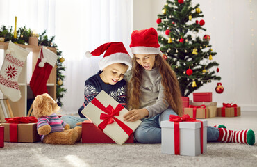 Christmas miracle. Cheerful little boy and girl in red Santa Claus hats happily open Christmas presents in morning. Kids look with interest at gift from which light shines and illuminates their faces.