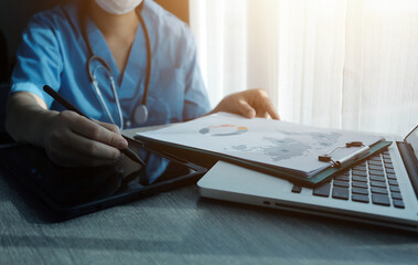 Doctor's working on laptop computer, writing prescription clipboard with record information paper folders on desk in hospital or clinic, Healthcare and medical concept. Focus on stethoscope