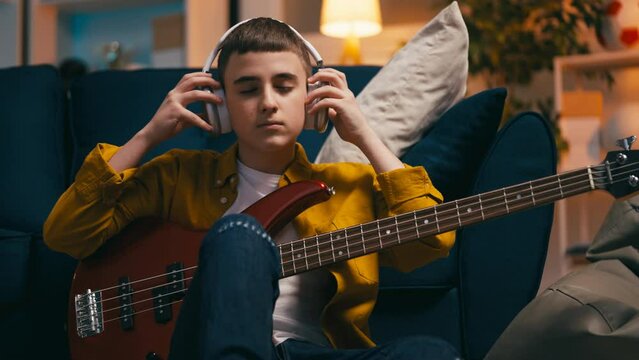 Teen boy putting headphones on and playing bass guitar at home, musical hobby
