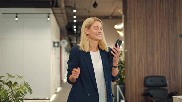Female financial director taking break from work and dancing at coworking space with mobile phone in hand. Cheerful fair-haired lady in elegant attire relaxing to cool music in empty corridor.